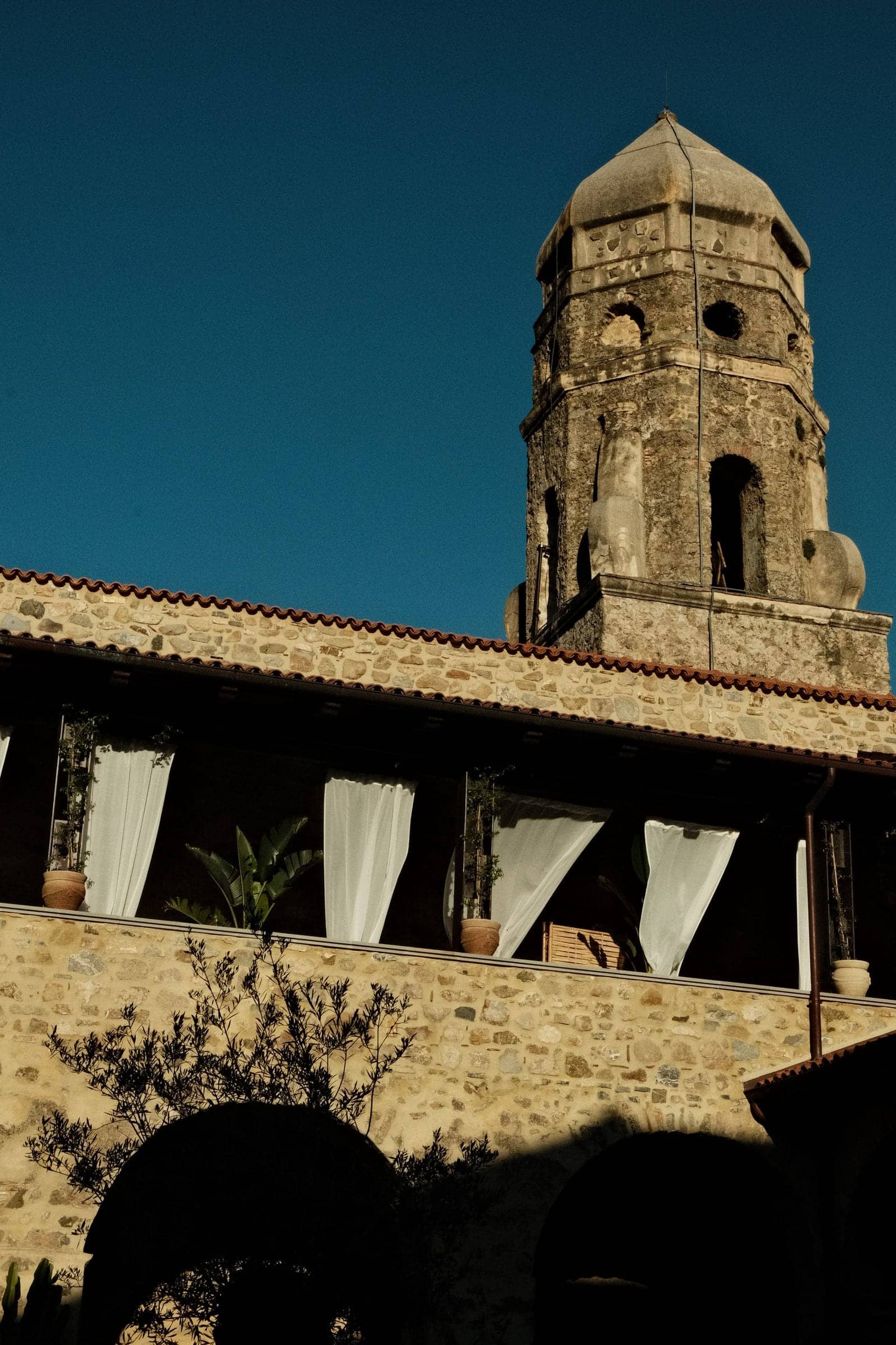 The stone bell tower of a former convent in the hills of the Cilento, white curtains lifting on the loggia below